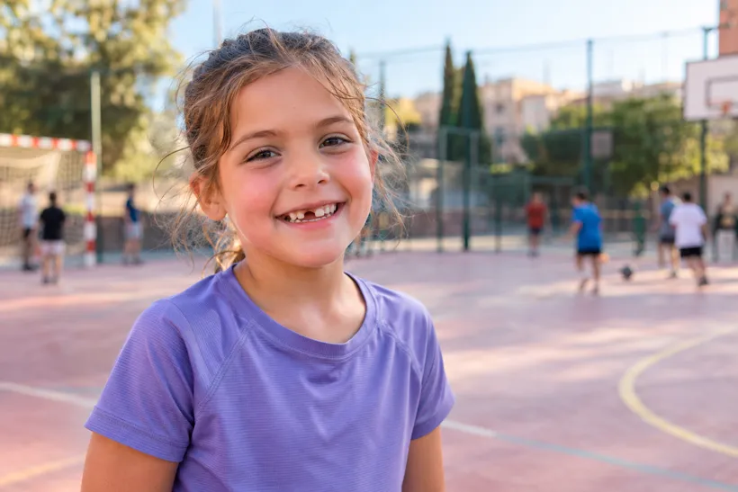 Niña con equipación deportiva sonriendo en una pista, con un diente de leche recién caído